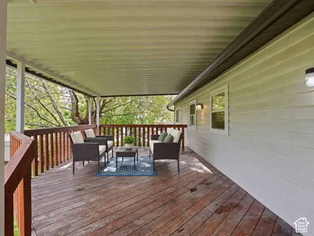 Covered deck featuring natural wood-finish plank flooring, a wooden railing, and exterior wall-mounted lighting