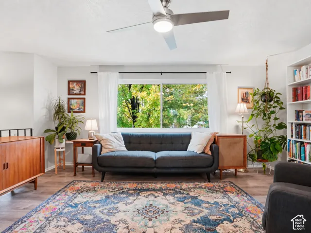 Living area featuring wood-finish flooring, a large horizontal window, brushed nickel ceiling fan with integrated lighting, and white built-in shelving