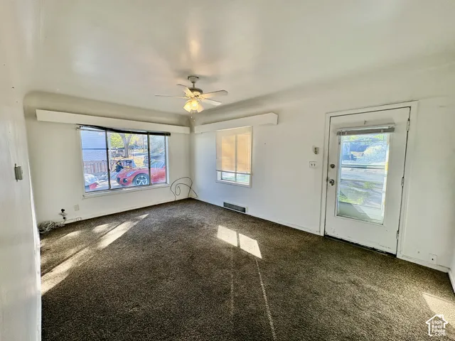Living area featuring large windows with box-style valances, a central ceiling fan with integrated lighting, carpeted flooring, and a glass-paneled exterior door