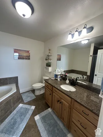 Bathroom featuring a dark stone-finish countertop, wood-finish cabinetry, an undermount oval sink with a dark-finish faucet, a tile-surround soaking tub, and wall-to-wall vanity mirror