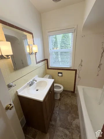Bathroom featuring a wood-finish vanity, integrated sink basin, chrome faucet hardware, tile wainscoting with accent trim, and neutral tile flooring