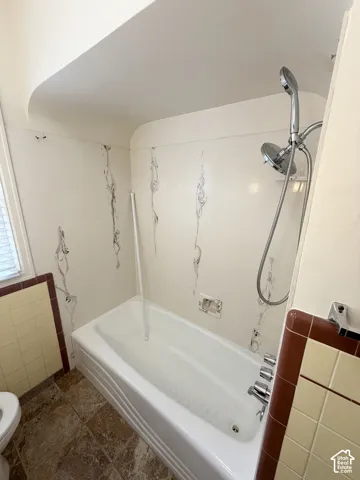 Bathtub enclosure with patterned wall surround, chrome handheld shower fixture, multi-handle faucet, and ceramic tile wainscoting