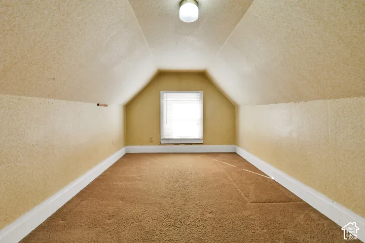 Finished attic space featuring vaulted ceilings, textured wall surfaces, neutral carpeting, and a single centered window