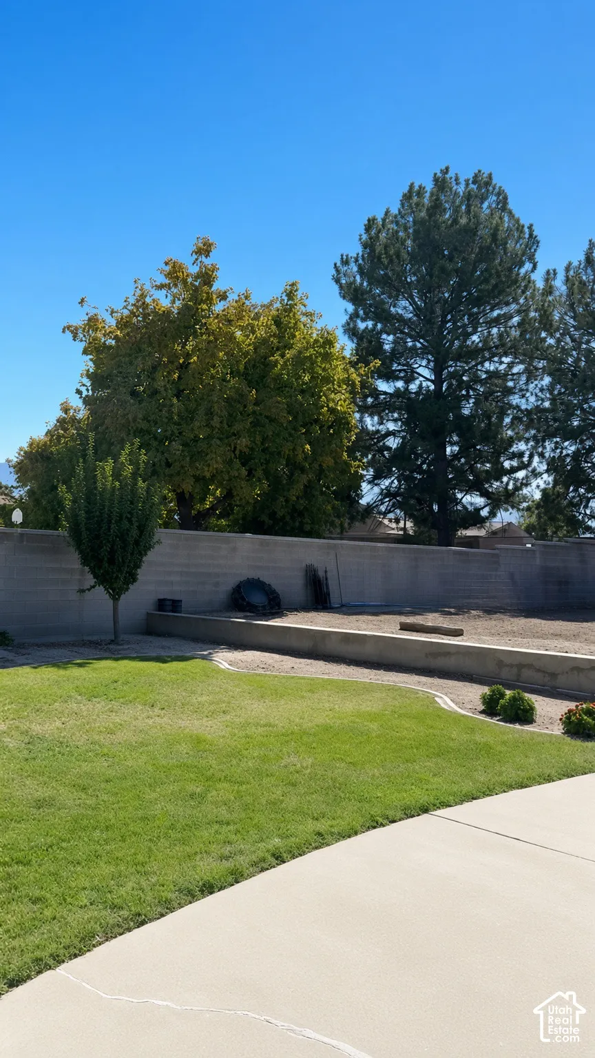 Backyard featuring a grass lawn, concrete patio, concrete block perimeter wall, terraced landscaping beds, and mature trees