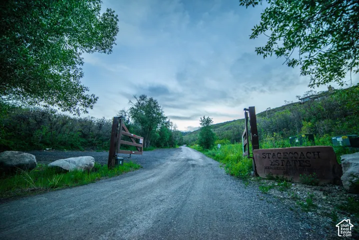 Gated entrance featuring a carved stone monument and heavy-duty timber gate, positioned at the start of a gravel access road lined with mature trees and natural hillside vegetation
