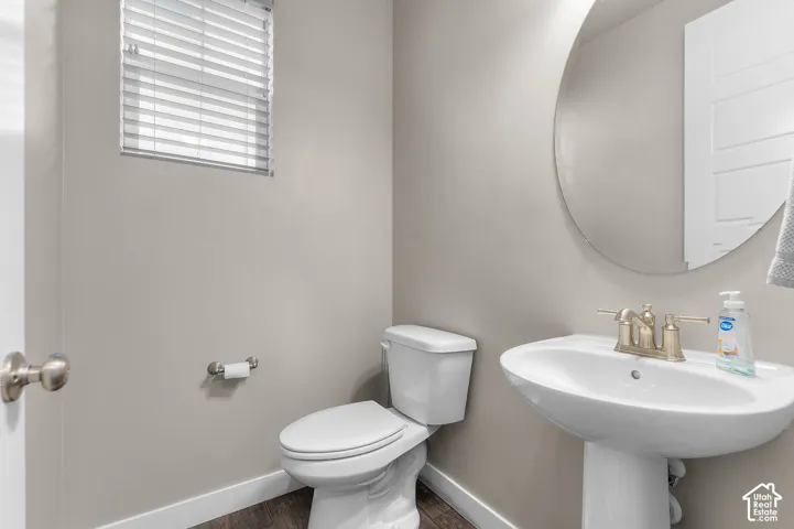 Pedestal sink with metallic faucet, frameless circular wall mirror, wood-finish flooring, and window with horizontal blinds