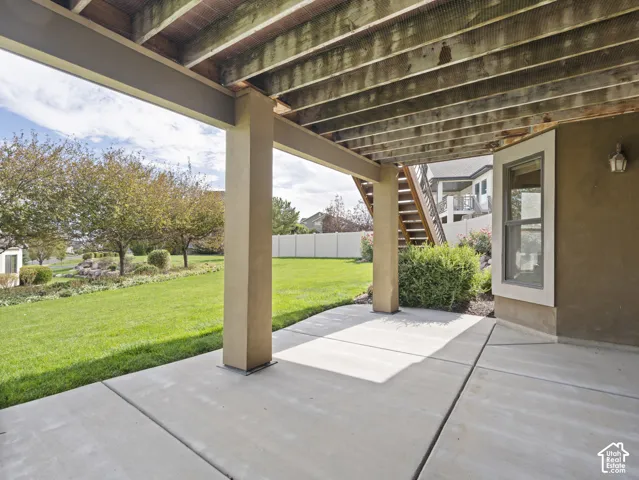 Covered concrete patio with structural support pillars, wood-beam ceiling, and integrated wall lighting, overlooking a manicured lawn and perimeter fencing