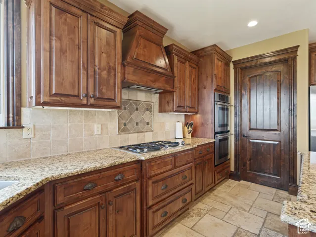 Kitchen featuring custom stained-wood cabinetry, stone tile flooring, integrated double wall ovens, gas cooktop with a matching wood vent hood, and stone countertops