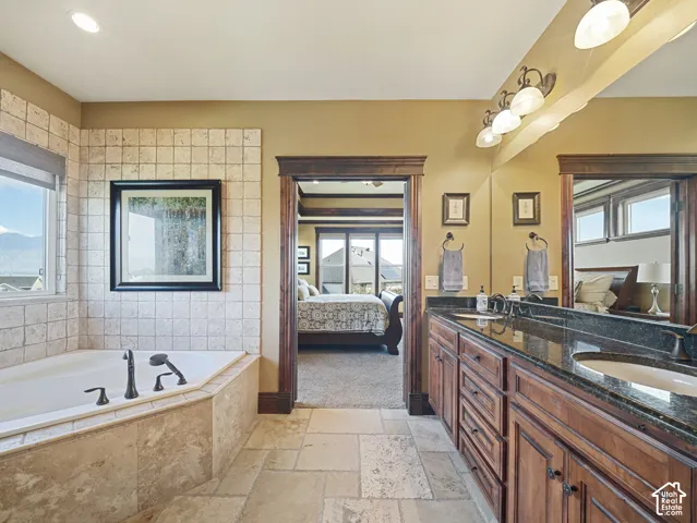 Deep soaking tub with stone tile surround, dual-sink vanity featuring dark stone countertops, natural stone flooring, and stained wood cabinetry with matching door casings