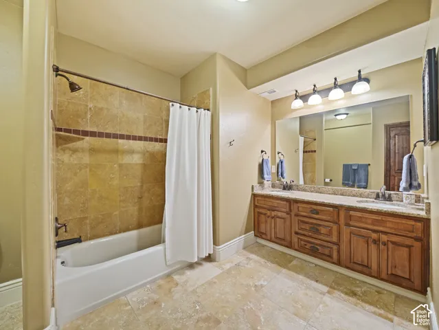 Bathroom featuring a dual-sink vanity with wood cabinetry and stone countertops, stone tile flooring, and a tub-shower combination with decorative stone tile surround