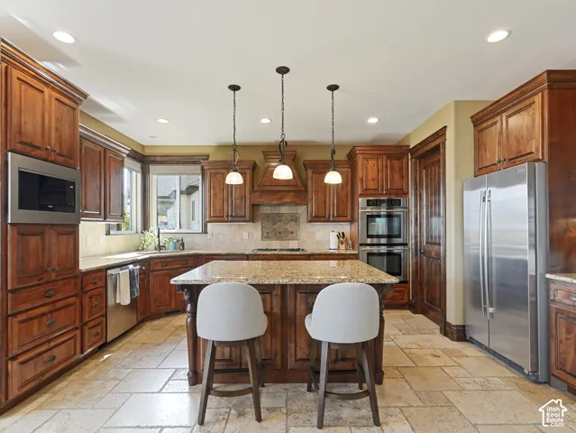 Kitchen featuring custom wood cabinetry, a central island with a stone countertop, tile flooring, stainless steel appliances, and decorative pendant lighting