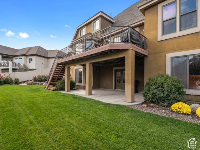 Stucco exterior featuring an elevated deck with wrought-iron railing, a covered ground-level patio, an exterior staircase, and a landscaped lawn