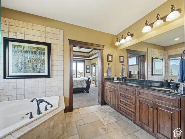 Dual-sink vanity with stone countertop and dark wood cabinetry, tile-surround soaking tub, wall-mounted light fixtures, and natural stone flooring
