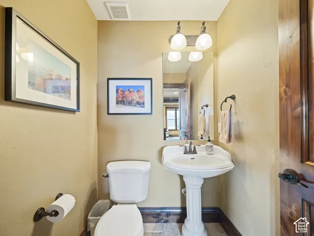 White pedestal sink featuring dual-handle faucet, wall-mounted mirror, two-light vanity fixture, and dark-toned wood-finish door with oil-rubbed bronze hardware