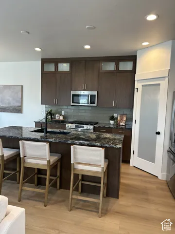 Kitchen featuring custom wood cabinetry with glass-panel upper accents, stone countertops, a central island with an integrated sink and matte black hardware, recessed ceiling lighting, and wood-finish flooring
