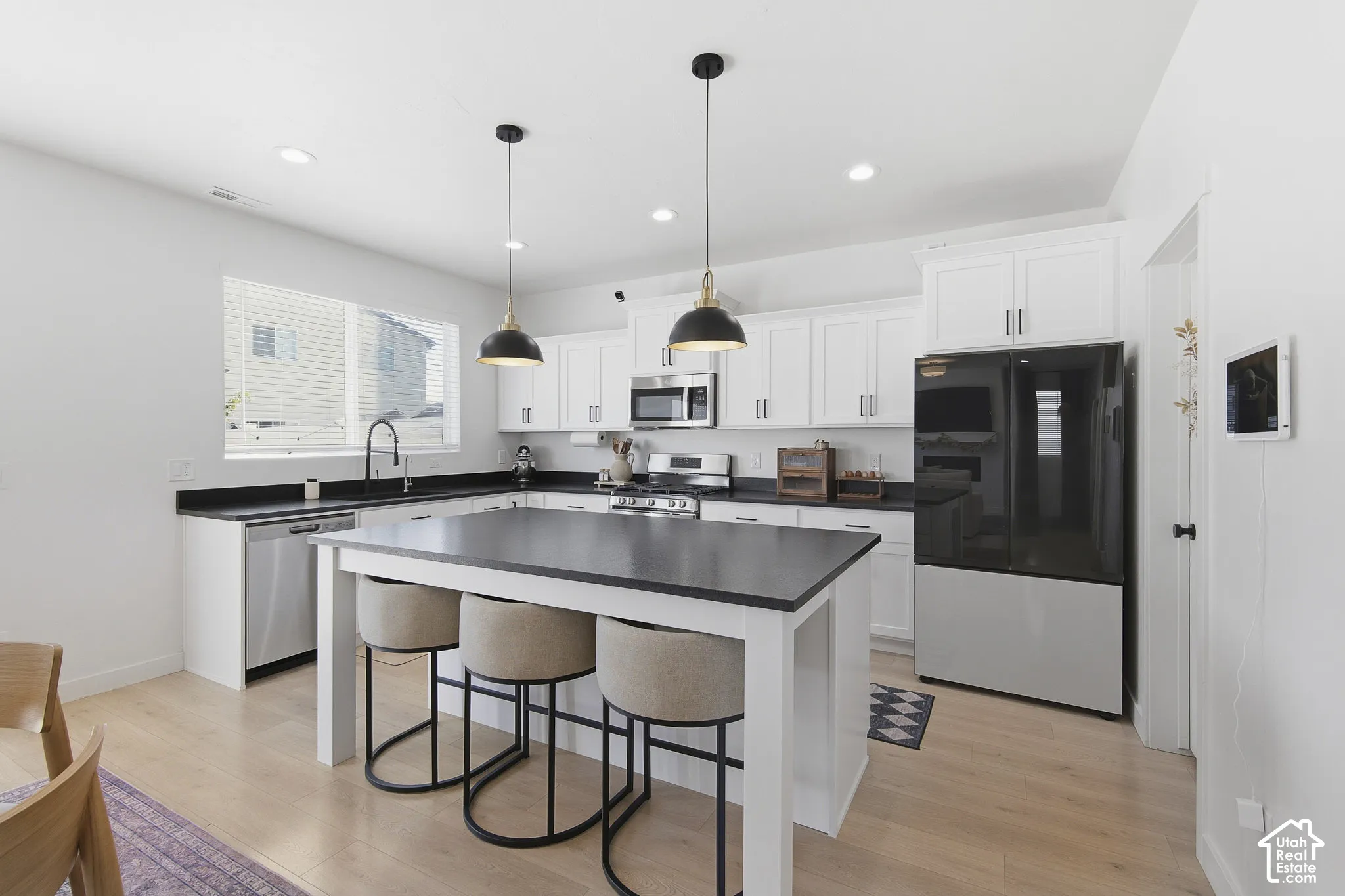 Kitchen featuring a central island with a contrasting countertop, white Shaker-style cabinetry, stainless steel appliances, matte black pendant lighting, and wood-finish flooring
