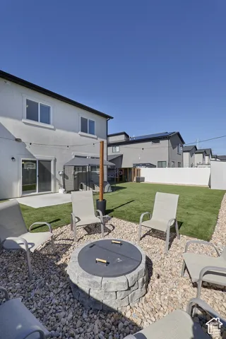 Backyard featuring a stone-block fire pit, gravel ground cover, artificial turf lawn, concrete patio, and white vinyl perimeter fencing