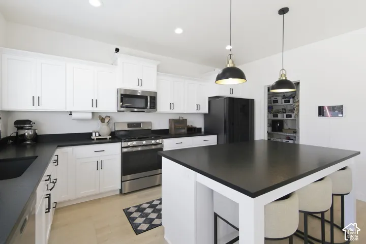 Kitchen featuring white shaker-style cabinetry, black countertops, stainless steel appliances, dual pendant lighting, and a kitchen island with built-in seating