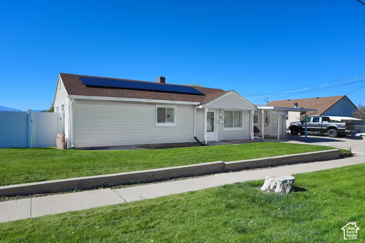 Single-story exterior featuring white horizontal siding, an asphalt shingle roof with integrated solar panels, a concrete driveway, a covered carport, and a white vinyl privacy fence