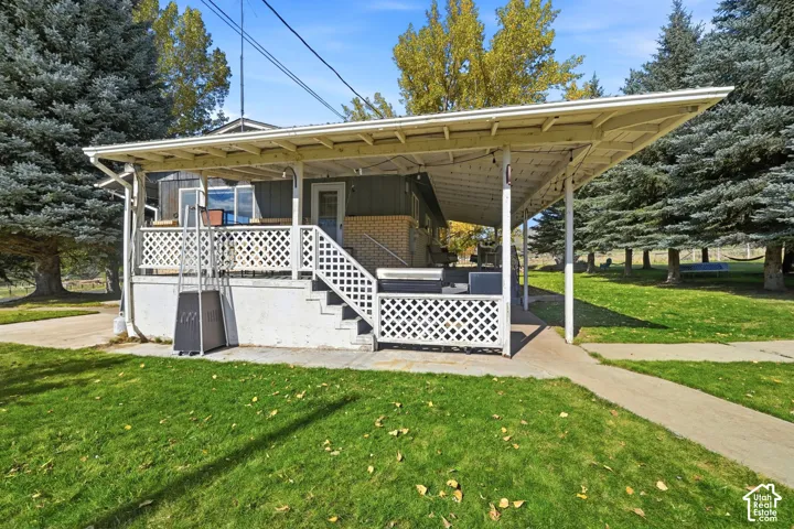 Covered patio featuring a concrete slab foundation, white lattice railings, and structural support posts, set within a grass-covered yard with mature evergreen trees