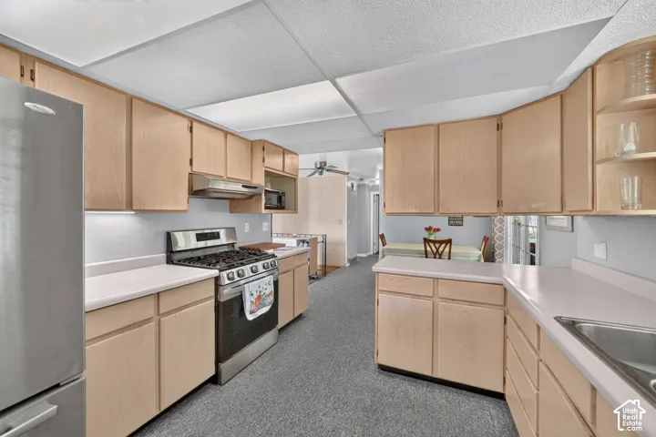 Kitchen featuring light-finish wood cabinetry, solid-surface countertops, stainless steel gas range, stainless steel range hood, and stainless steel refrigerator