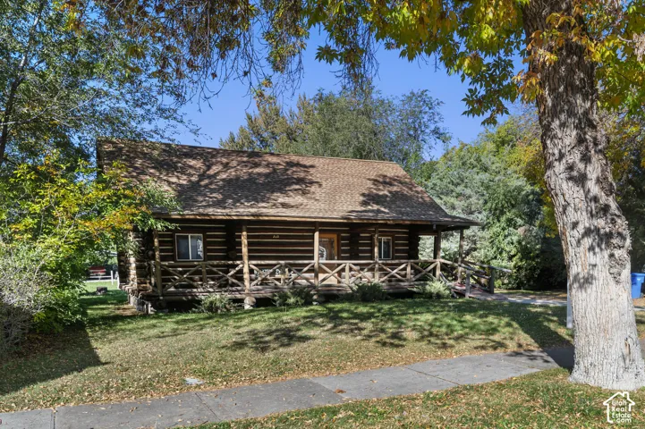 Log exterior home featuring a covered front porch with a wood railing and shingled roof, surrounded by mature trees