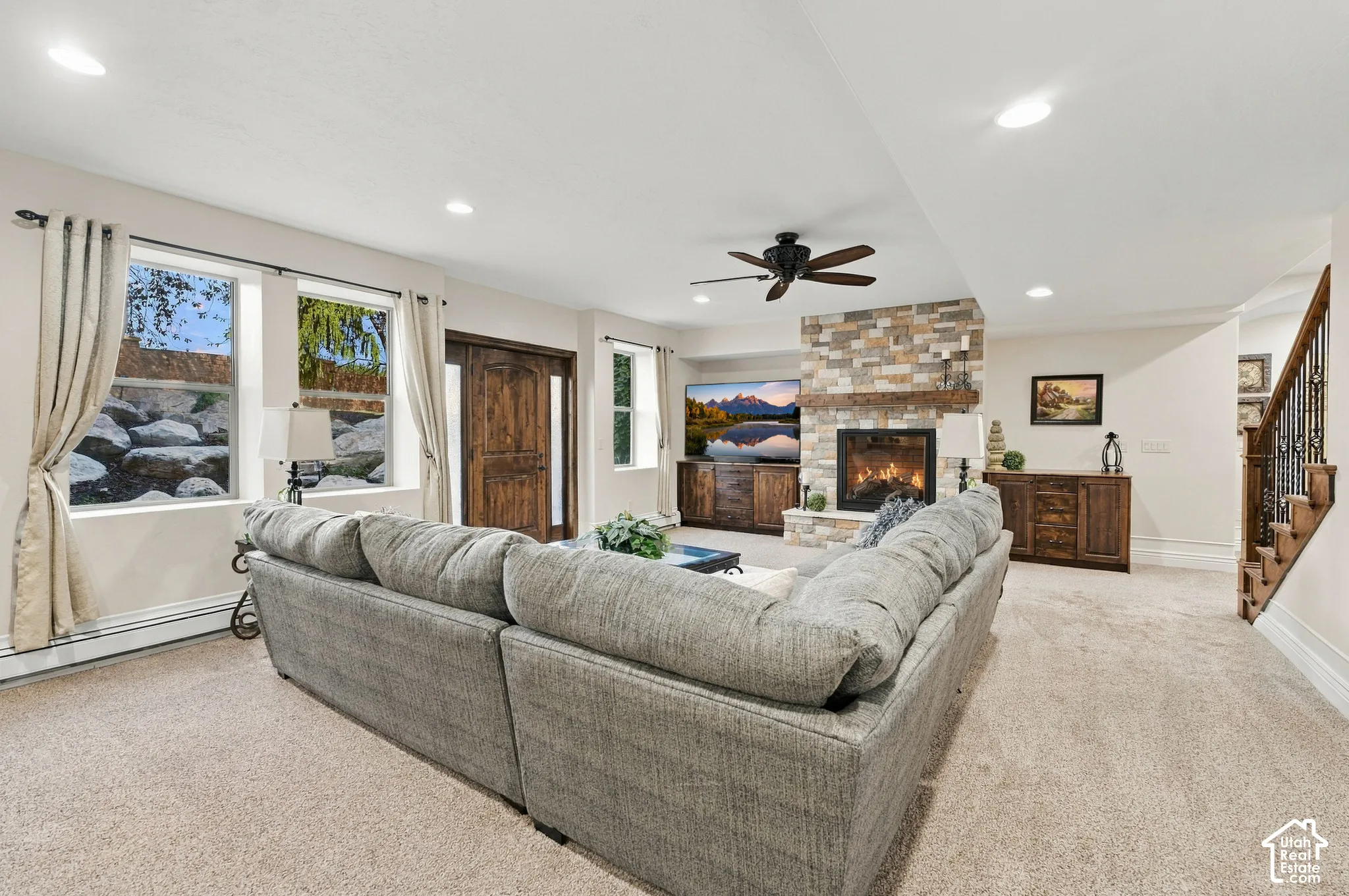 Full-height stone fireplace with timber mantel, built-in cabinetry, recessed ceiling lighting, ceiling fan, and staircase with metal balusters.