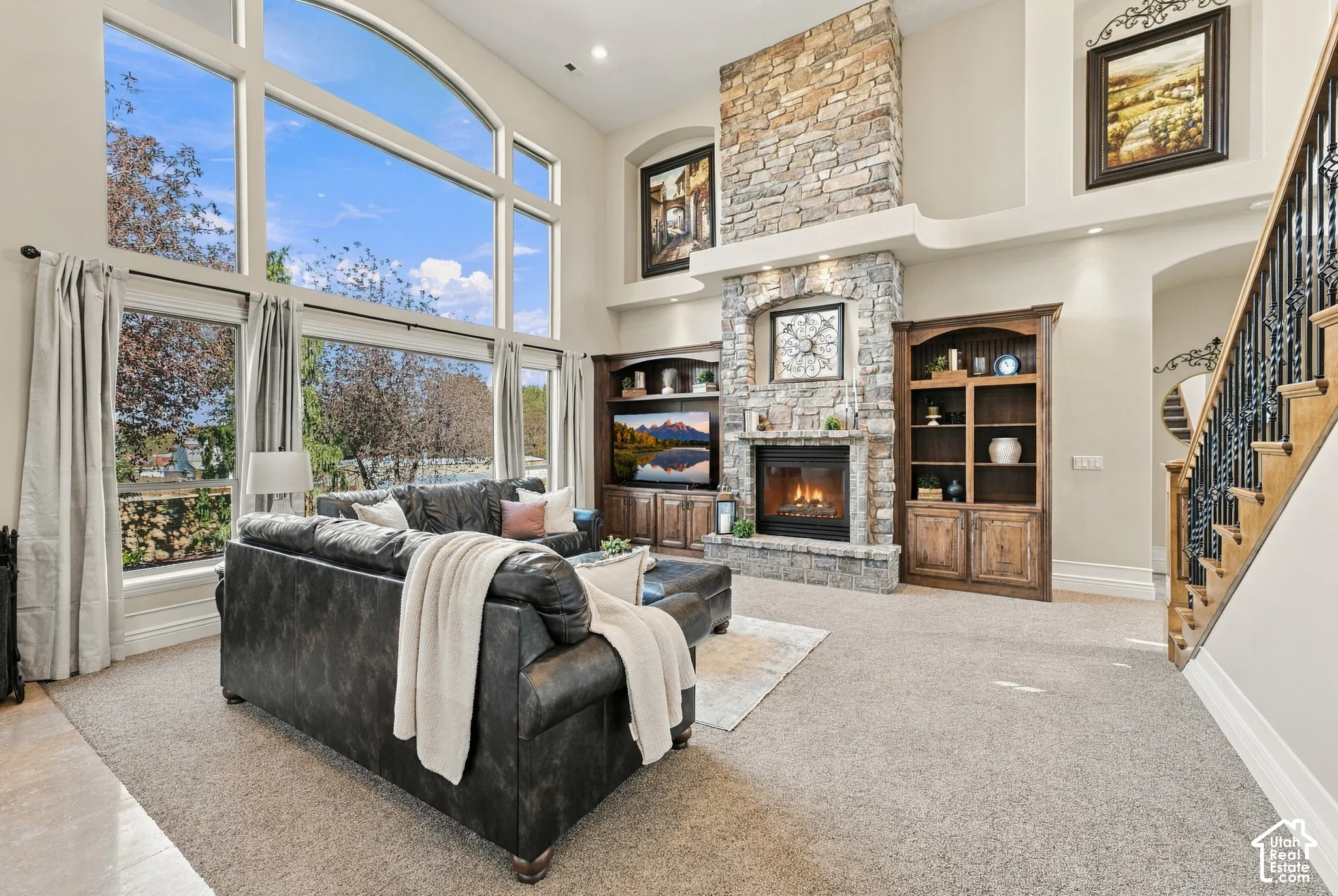 Double-height living area featuring floor-to-ceiling windows, a floor-to-ceiling stone fireplace, built-in wooden bookcases, and a wooden staircase with decorative metal railing.