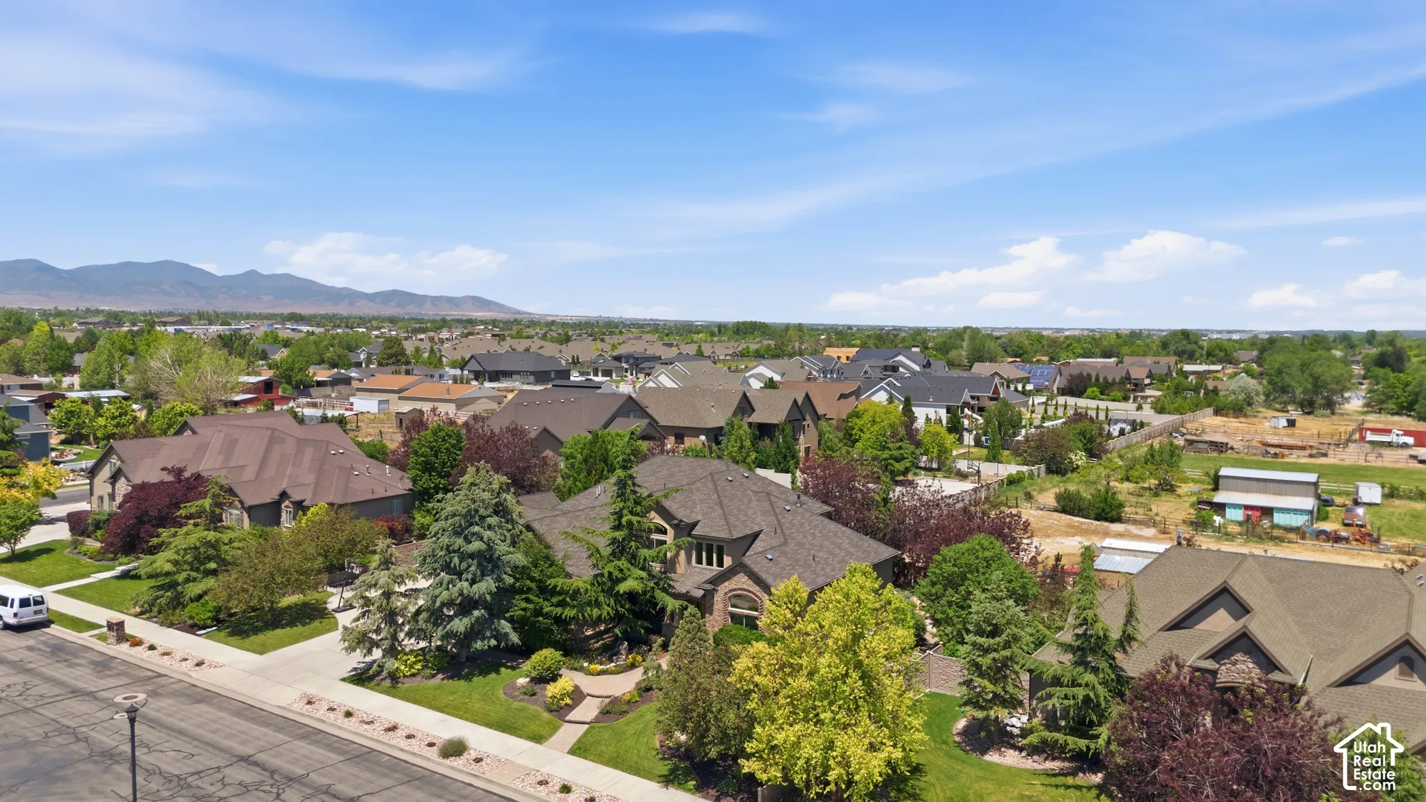 Aerial perspective featuring a residential property with a multi-gable roofline, paved driveway, landscaped grounds with mature evergreen trees, and a mountain horizon backdrop.