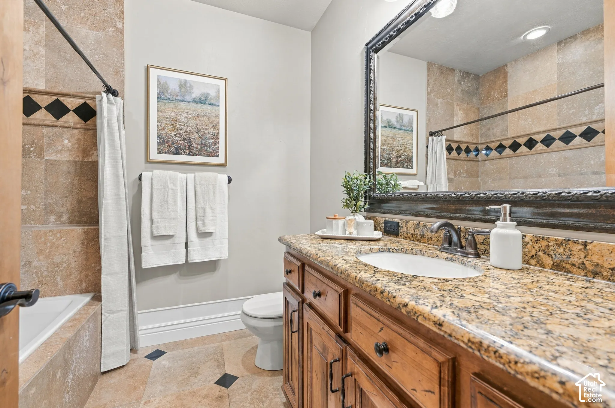 Bathroom featuring a wood vanity with stone countertop, drop-in sink, large framed mirror, and tiled shower surround with decorative diamond-pattern inlay.