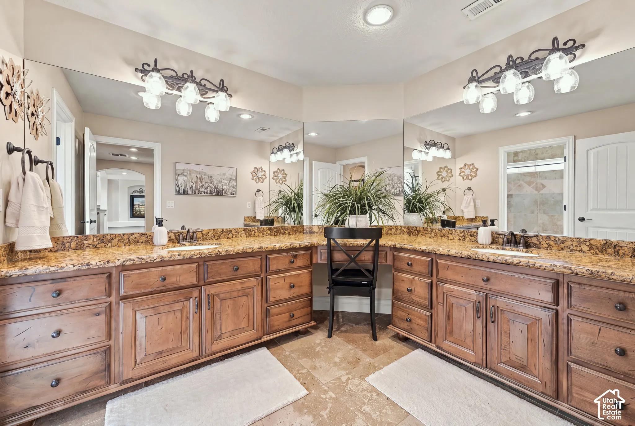 Dual-sink vanity with stained wood cabinetry, stone-surface countertops, integrated sit-down makeup desk, and multi-panel wall mirror.