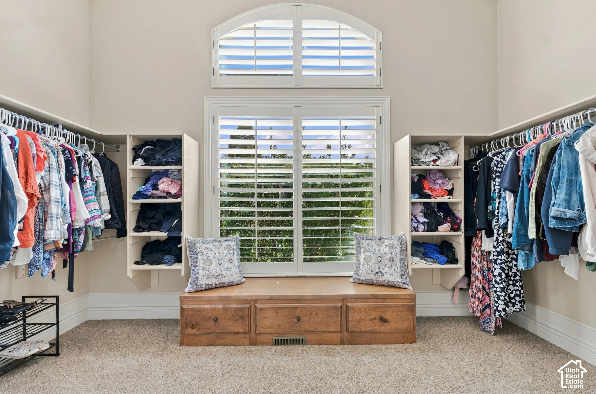 Walk-in closet featuring built-in shelving, double-tier clothing rods, a wood-finish storage bench with drawers, and an arched window with plantation shutters.