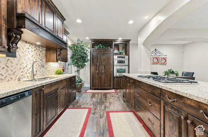 Kitchen featuring custom dark wood cabinetry with decorative corbels, natural stone countertops, a mosaic tile backsplash, wood-finish flooring, and integrated stainless steel appliances