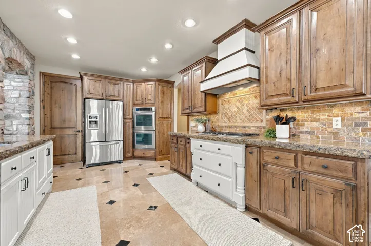 Kitchen featuring custom wood cabinetry, stone tile flooring with decorative inserts, a natural stone backsplash, stainless steel double wall ovens, and a custom range hood.