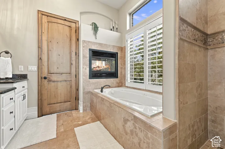 Built-in soaking tub with a tiled surround, integrated fireplace, plantation shutters, stone-tile flooring, and solid wood-paneled door.