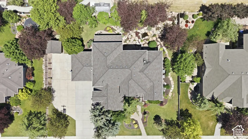 Aerial perspective featuring a multi-hipped asphalt shingle roof, expansive concrete driveway, stone-accented landscape bed, mature trees, and a paved walkway.