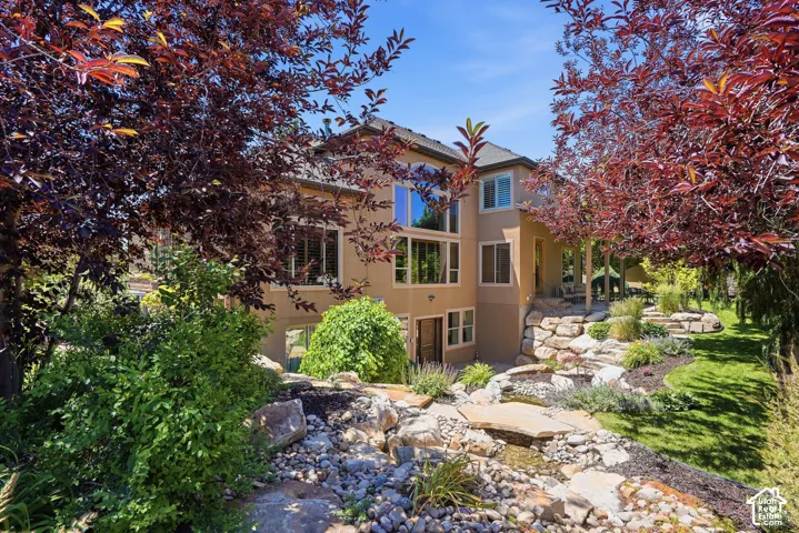 Stucco exterior featuring multi-story windows, stone retaining walls, rock landscaping, mature trees, and a tiered lawn.
