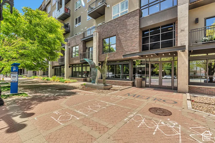 Multi-story exterior featuring a brick facade, grid-pattern windows, private balconies, and a paved pedestrian plaza with sculptural elements