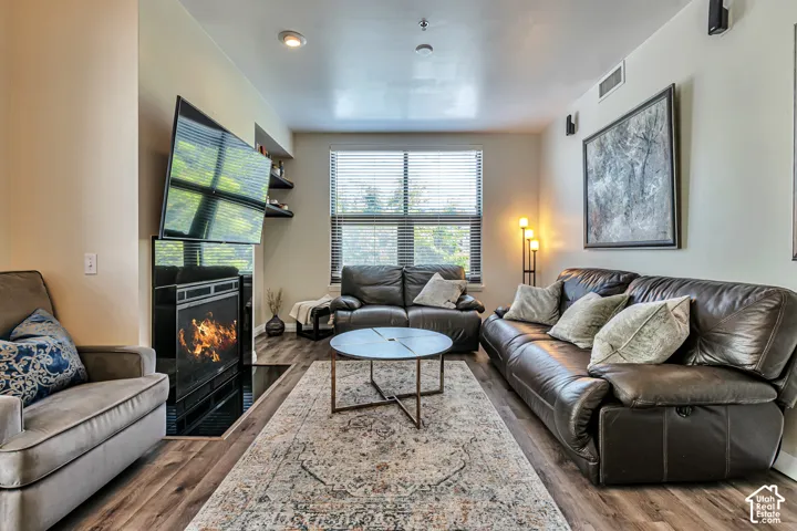 Living area featuring wood-finish flooring, a built-in fireplace with a black glass surround, recessed lighting, and a large window with horizontal blinds