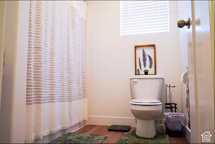 Bathroom featuring wood-finish flooring, a white porcelain toilet, and a window fitted with horizontal blinds