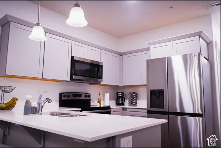 Kitchen featuring light-gray Shaker-style cabinetry, white solid-surface countertops, stainless steel appliances, dual pendant lighting, and a dual-basin undermount sink