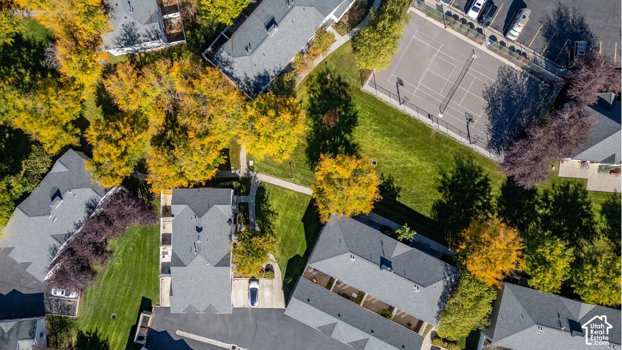 Aerial perspective of the property featuring asphalt shingle roofing, paved driveways, a manicured lawn, mature trees, and a hard-court tennis surface