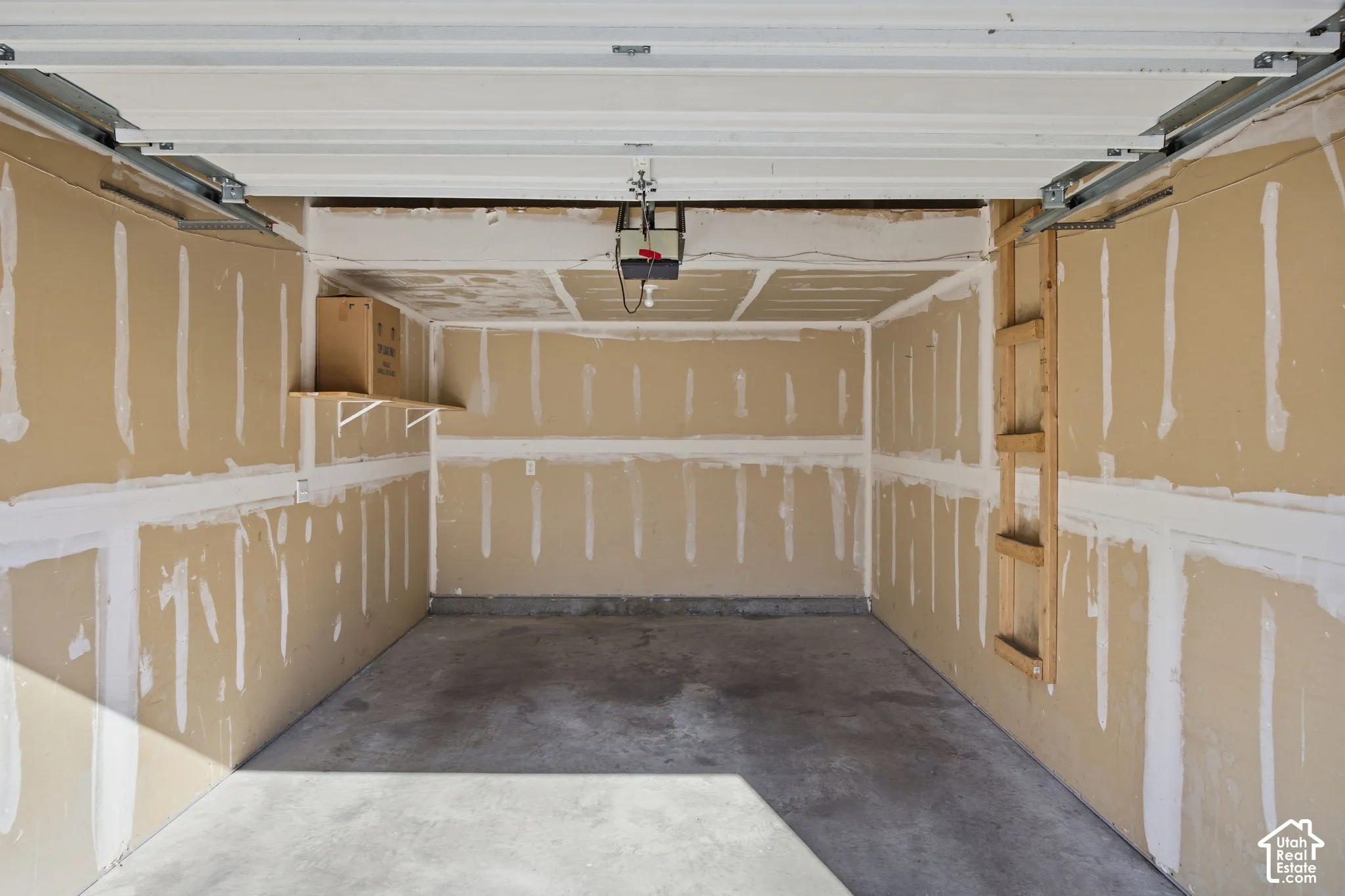 Garage featuring concrete flooring, an automatic overhead door mechanism, unfinished drywall walls, and a built-in wall-mounted wooden shelf