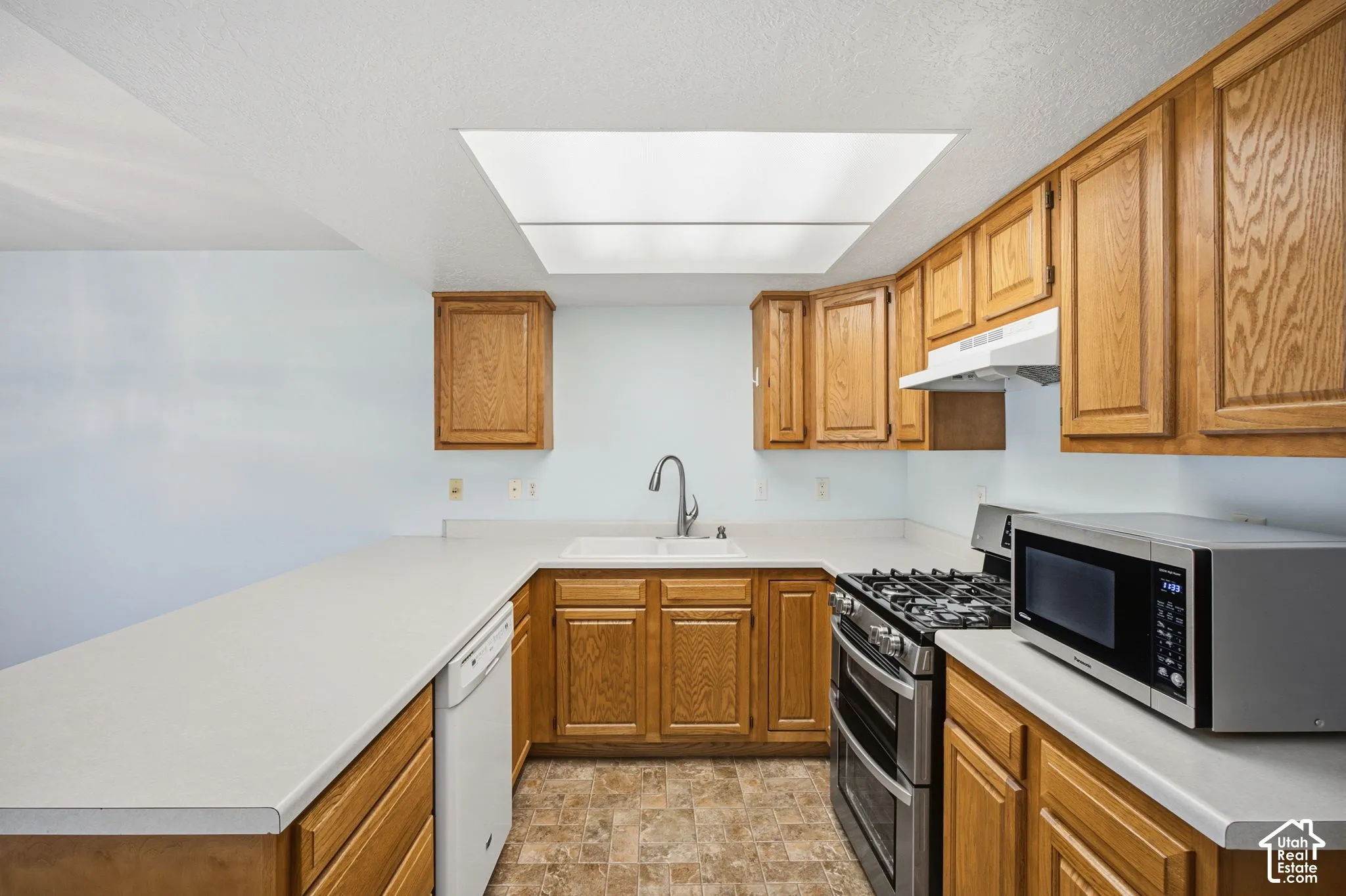 Kitchen featuring natural wood cabinetry, solid-surface countertops, dual-basin sink with arched faucet, stainless steel gas range, and recessed ceiling lighting