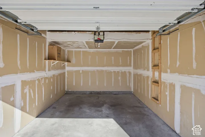 Garage featuring concrete flooring, an automatic overhead door mechanism, unfinished drywall walls, and a built-in wall-mounted wooden shelf