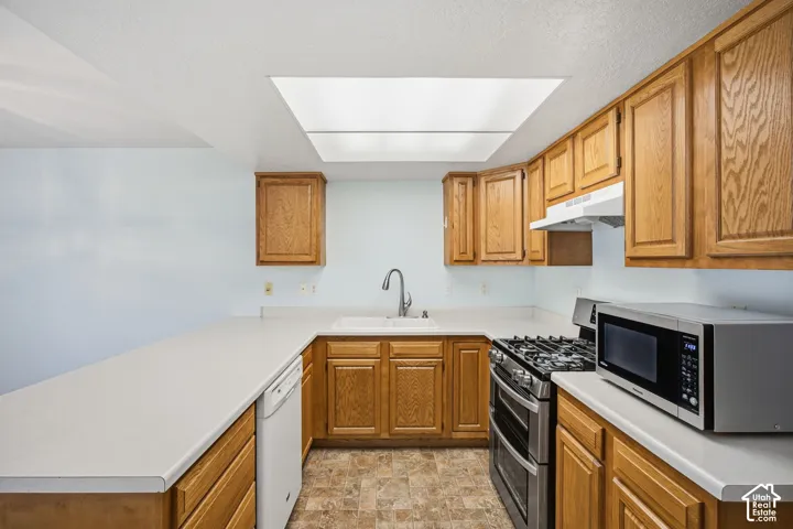Kitchen featuring natural wood cabinetry, solid-surface countertops, dual-basin sink with arched faucet, stainless steel gas range, and recessed ceiling lighting