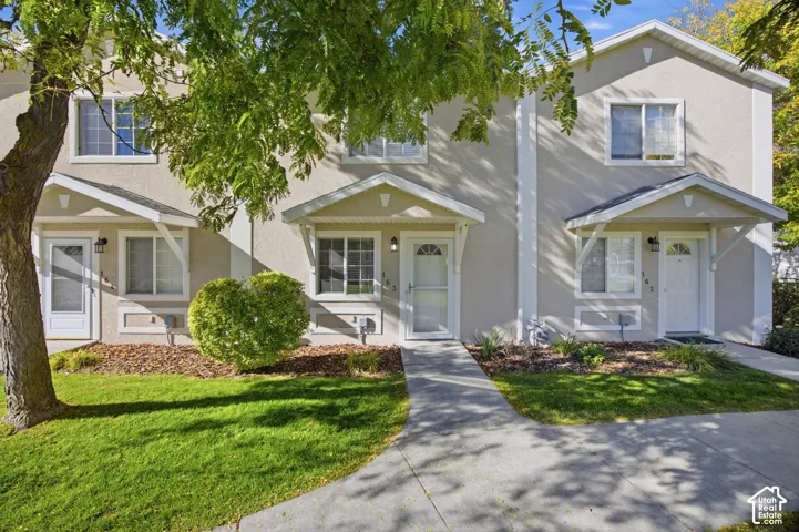 Two-story townhouse exterior featuring light-toned stucco siding, gabled roofline entryways, concrete walkways, and landscape-integrated planting beds