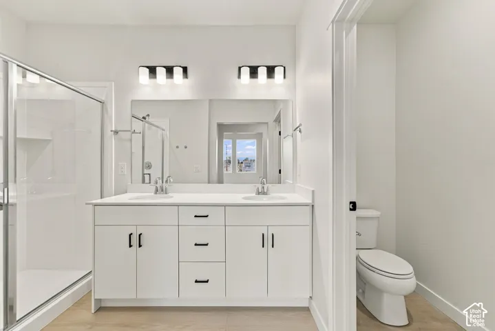 Dual-sink vanity with white cabinetry, quartz countertop, glass-enclosed shower, wood-finish flooring, and wall-mounted vanity lighting