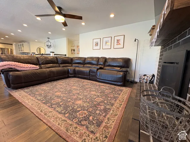 Dark-stained wood-finish flooring, recessed lighting, ceiling fan with integrated lighting, and a fireplace featuring a brick surround and a prominent wood mantel