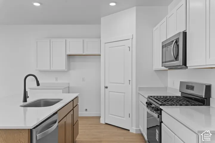 Kitchen featuring white shaker-style cabinetry, solid-surface countertops, a center island with an undermount sink and matte black pull-down faucet, stainless steel appliances, and wood-finish flooring
